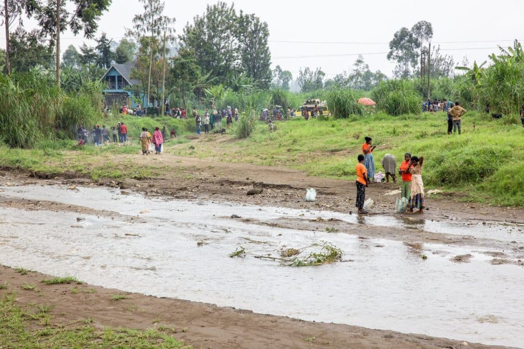 People gather near a small stream in a grassy area with scattered trees and distant houses in the background.