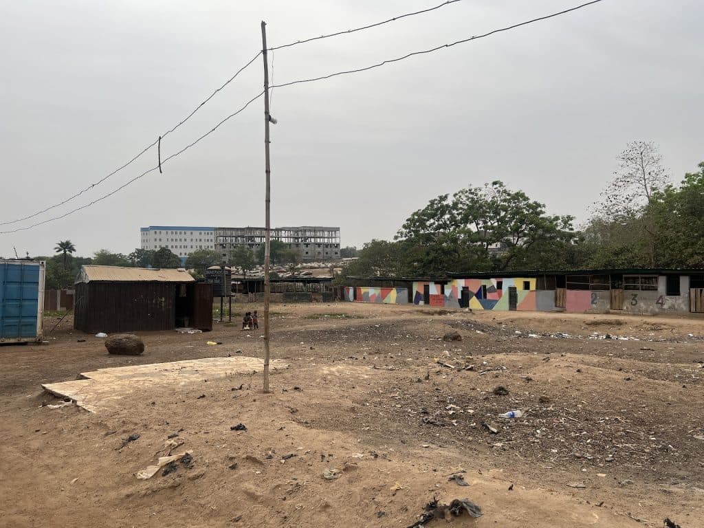 Open dirt area with scattered trash, a few small buildings, colorful wall murals, and sparse trees under a cloudy sky.