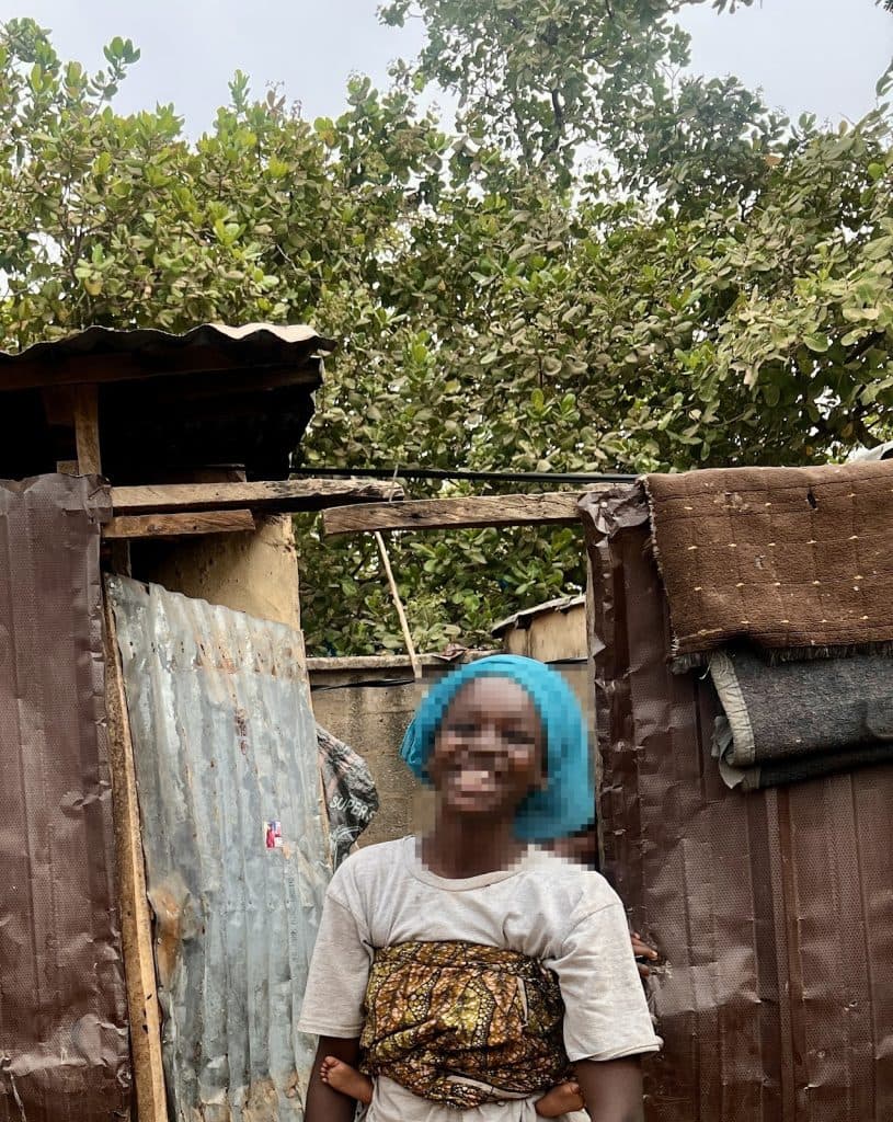 Smiling person with baby in a wrap stands near a makeshift structure, surrounded by green trees.