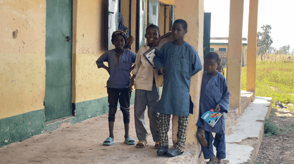 Four children stand on a porch, two are holding notebooks. A building and grassy field are in the background.