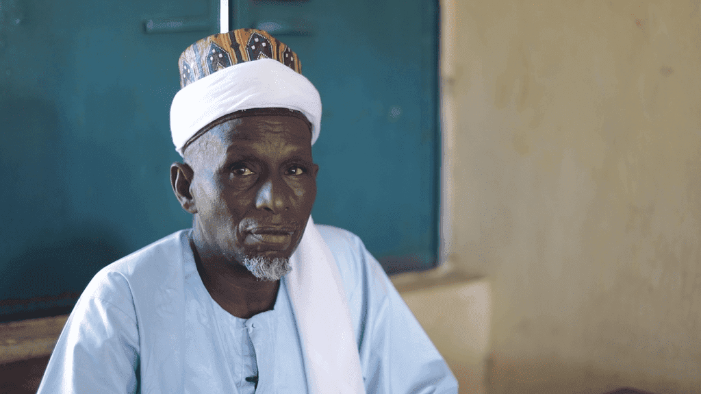 Elderly man in traditional attire, wearing a patterned hat and white headscarf, seated indoors with a blue door in the background.