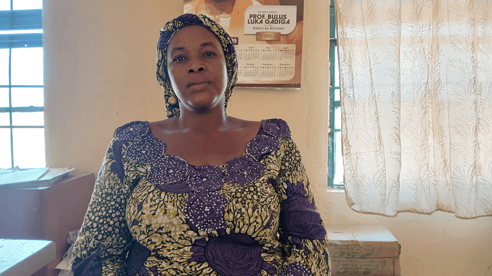 A woman in a colorful headwrap and dress sits indoors, with a calendar and window in the background.