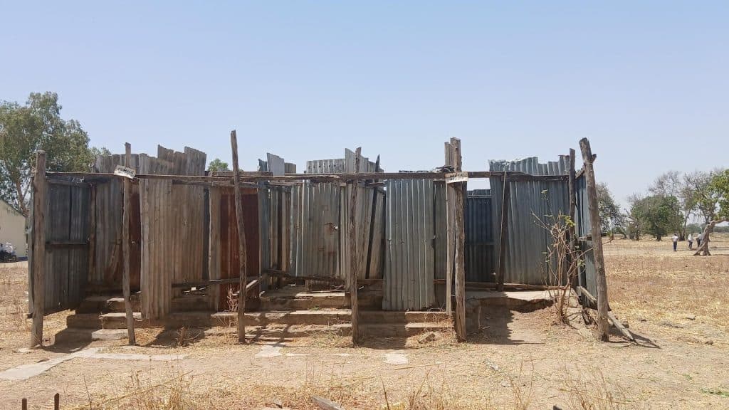 Rustic corrugated metal structure in an open field under a clear blue sky. Trees and people visible in the distant background.