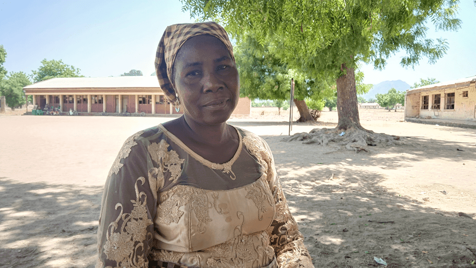 Woman in traditional attire stands under a tree, with a school building and blue sky in the background.
