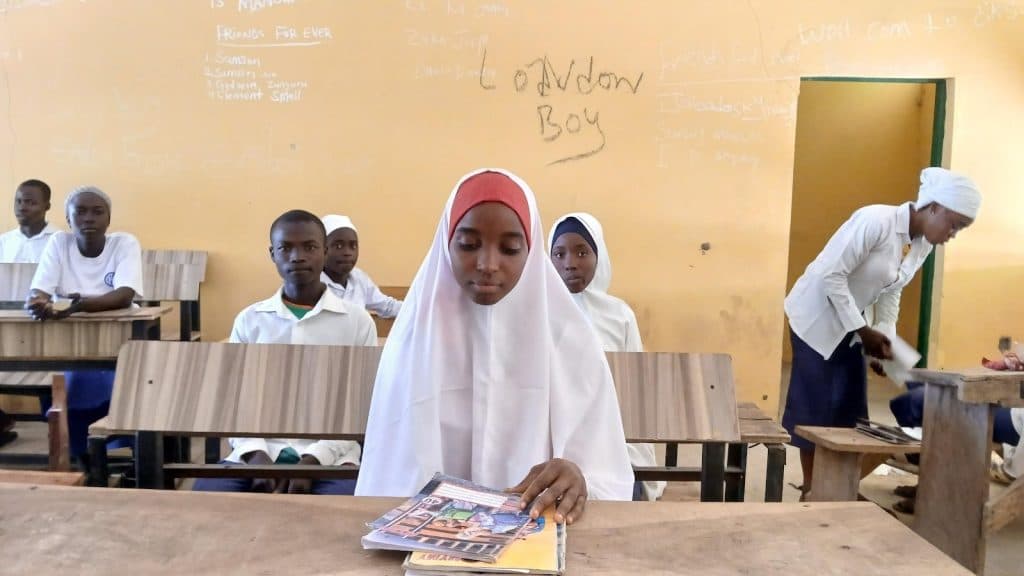 Students in a classroom with wooden desks, one girl in the front looks at her notebooks. Writing is visible on the chalkboard wall.