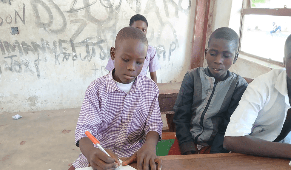 A boy in a checkered shirt writes with a pen, sitting at a wooden desk with other students in a classroom with graffiti on the wall.