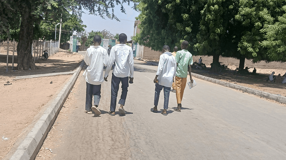 Four people walk down a sunny tree-lined street, two in white shirts and two in colored pants, carrying items in their hands.