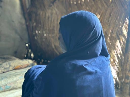 A person in a blue hijab sits indoors with a straw wall backdrop, looking away from the camera.