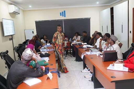 A person presenting to a diverse group in a conference room, with participants seated at a U-shaped table, engaged and taking notes.
