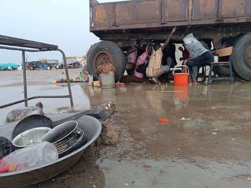 People seek shelter under a truck during a rainstorm. Nearby, a basin with dishes and bottles sits on the muddy ground.