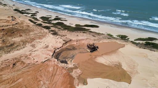 Aerial view of a beach with a large pit and a machine beside the ocean, surrounded by sand dunes and sparse vegetation.