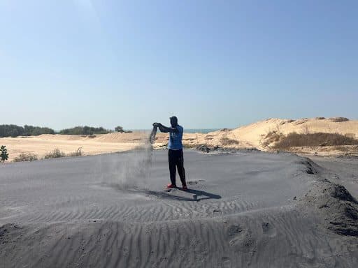 Person pours sand through their hands on a dark sandy area, with dunes and clear blue sky in the background.