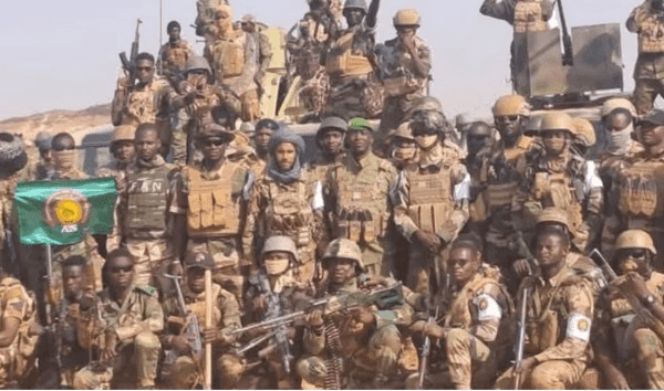 A large group of soldiers in camouflage uniforms with helmets and gear pose together outdoors, some holding weapons and a green flag.