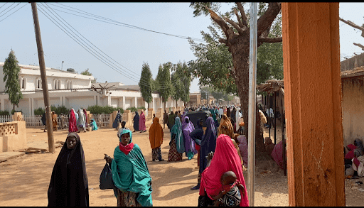 People in colorful clothing gather on a street lined with trees and houses, under clear skies.