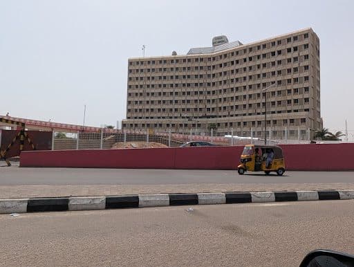 A yellow auto rickshaw drives past a large, multi-story building and a red wall on a clear day.