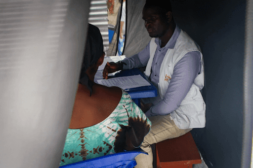 A healthcare worker in a white vest consults with a person seated across a table inside a tent, with papers on the table.