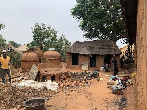 A rural scene with a thatched-roof house, clay structures, scattered items, and two people outside, surrounded by trees and dirt ground.