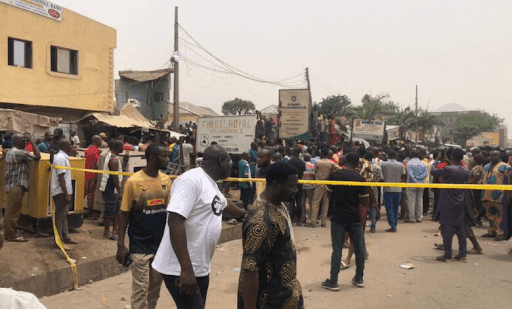 Crowd gathered in a street enclosed by yellow tape, with buildings and signs in the background.