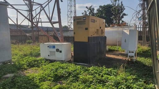 Outdoor generators and equipment surrounded by greenery near a metal tower.