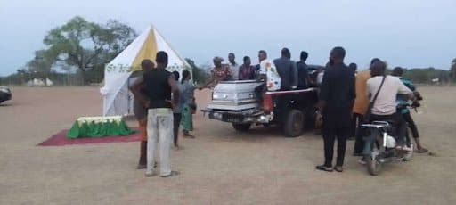 A group of people gather around a tent and a pickup truck carrying a coffin on a field, with a motorcycle nearby.