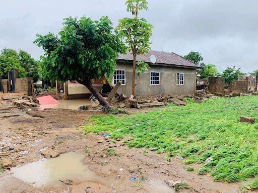 A small house surrounded by floodwater, with a muddy yard and green plants in the foreground, under a cloudy sky.
