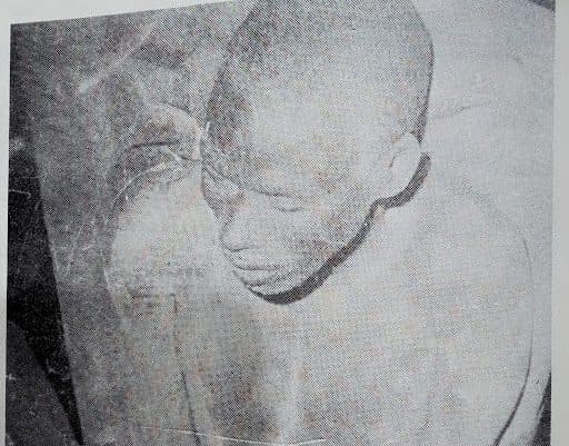 A grainy black-and-white photo of a person with a shaved head looking down, appearing seated or crouched.