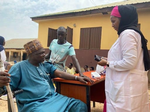Healthcare worker checks a man's blood pressure at an outdoor clinic, as another man assists, with people in the background.