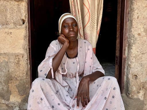 A woman in patterned robes sits in a doorway, resting her chin on her hand, with a thoughtful expression.
