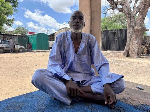 A man in a loose, light blue outfit sits cross-legged on a blue mat outdoors, with trees and buildings in the background.