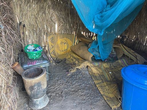 Inside a thatched hut, a bed with a blue mosquito net, a basket on a stool, a large pestle, and a blue container are visible.