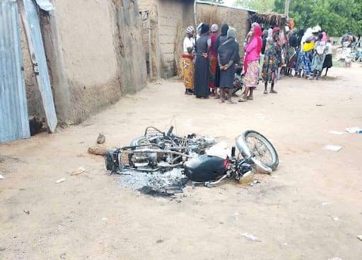 Burned motorcycle on sandy street, group of people in colorful clothing gathered near buildings in the background.