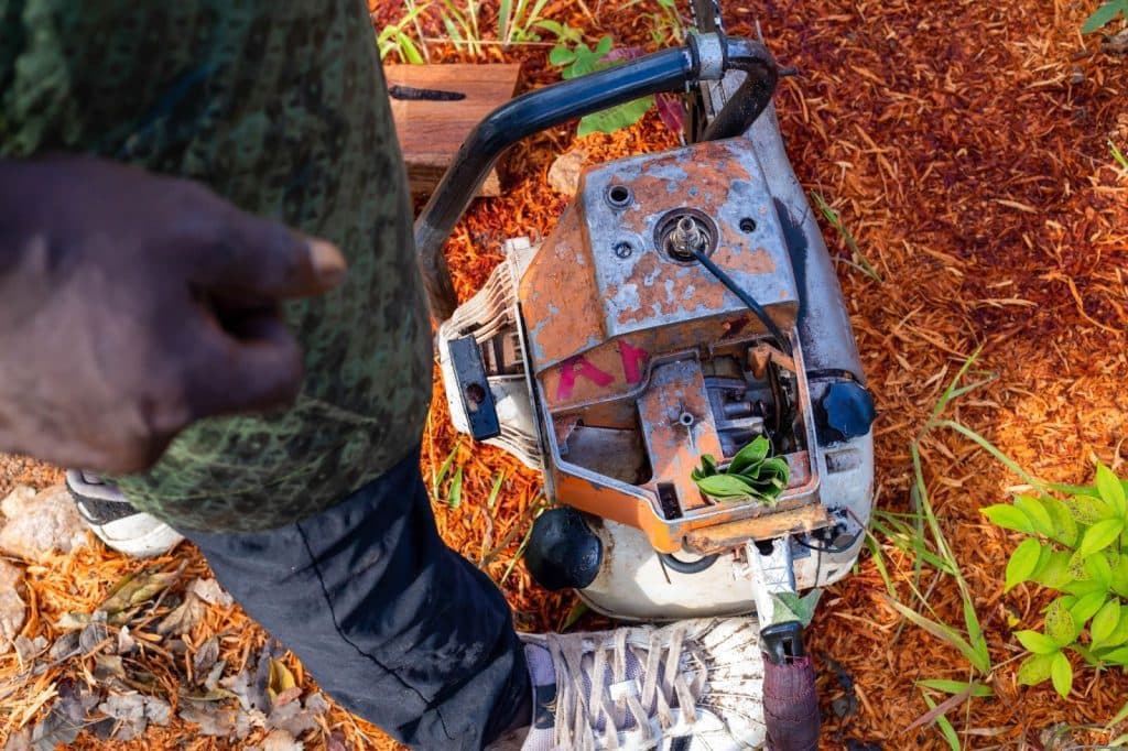 A person stands on a chainsaw lying on wood chips, with leaves stuck in its engine.