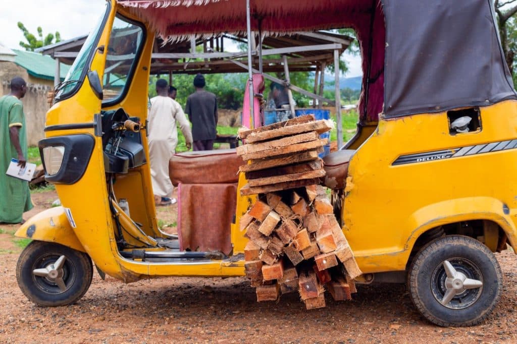 A yellow tricycle loaded with wooden planks parked on a dirt road, with people in the background.