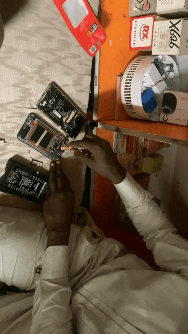 Person repairing a smartphone with disassembled parts on a table, surrounded by tech gadgets and boxes.