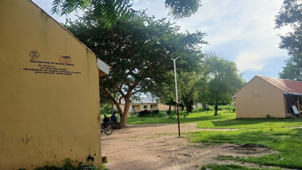 Buildings with "Construction of Market Shade" text, trees, and people sitting, with grass and a clear sky in the background.