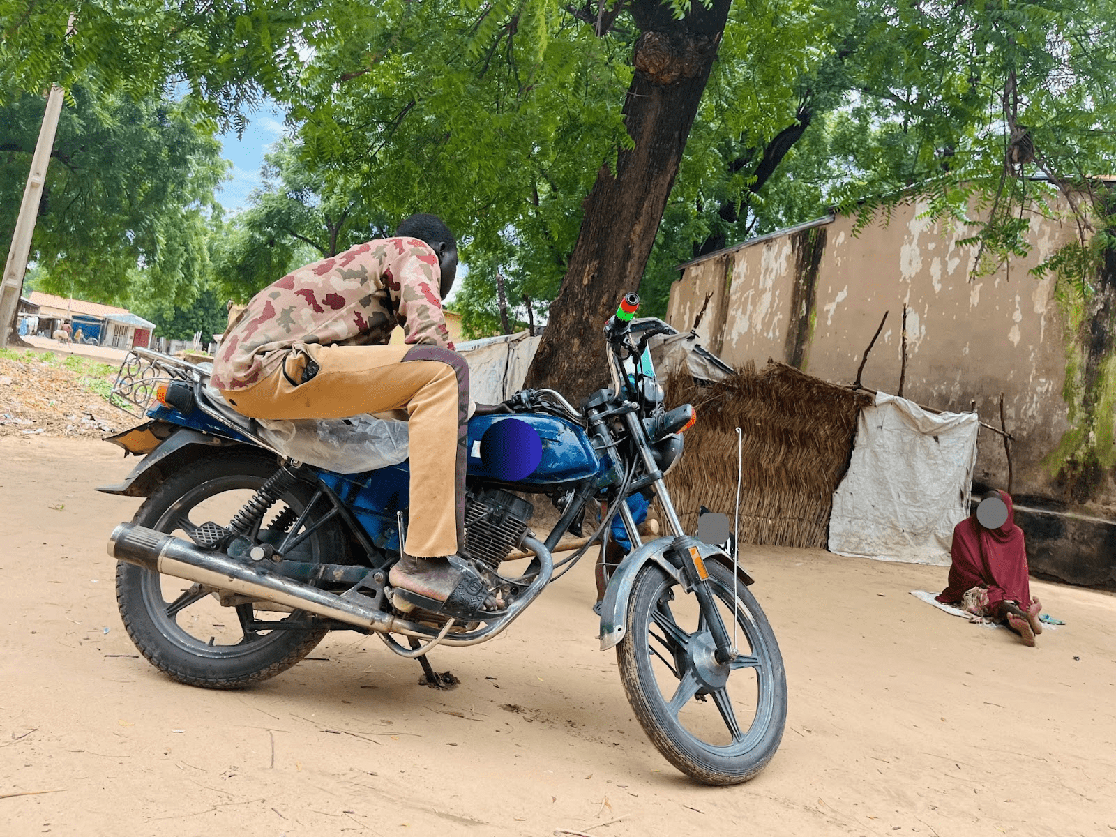 Person on a motorcycle beside a tree, with another person sitting on the ground nearby, against a backdrop of a rustic building and huts.