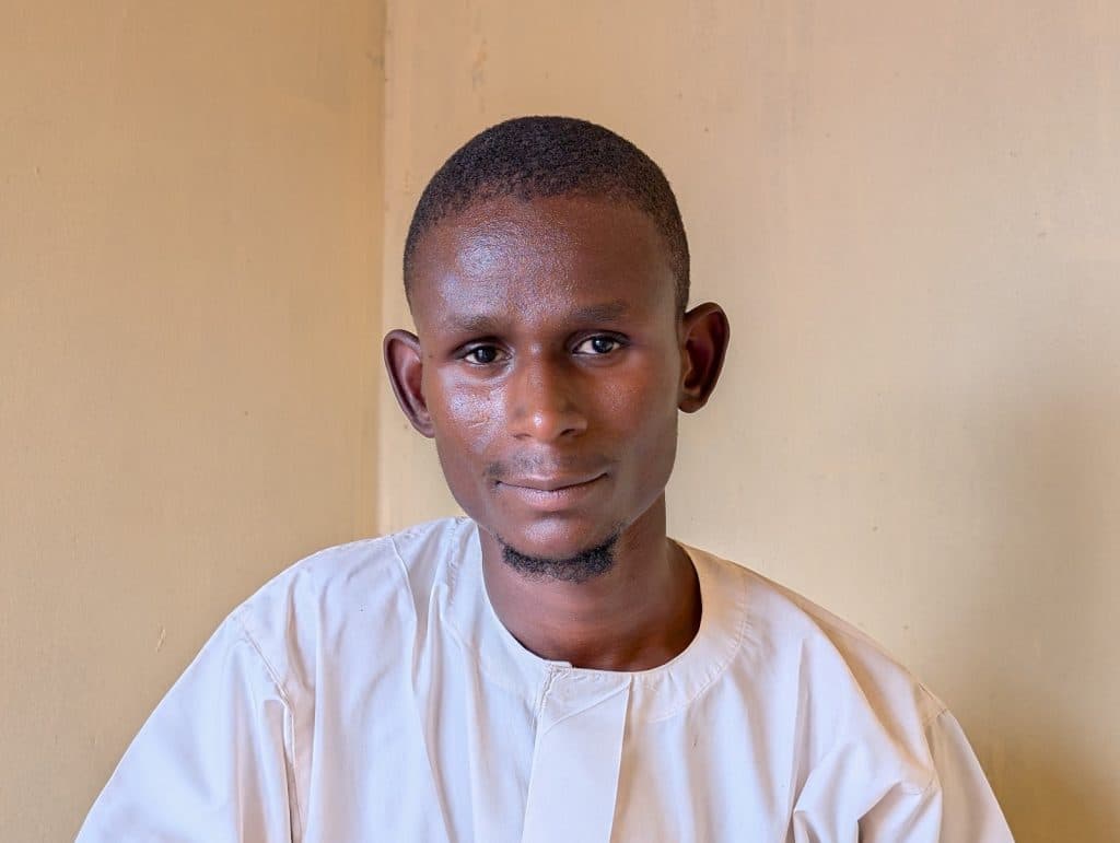 Man in a light-colored shirt stands against a plain wall, looking at the camera with a neutral expression.