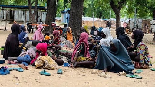A group of people wearing colorful clothing sit on the ground under trees, in an outdoor setting with scattered sandals nearby.