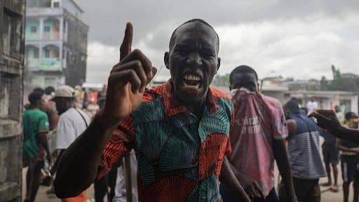 Man in colorful shirt passionately gestures amidst a crowded street scene, under an overcast sky.