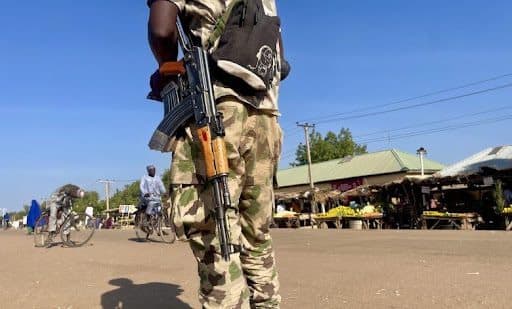 Armed soldier in camouflage patrols a busy street with bicycles passing by, under a clear blue sky.
