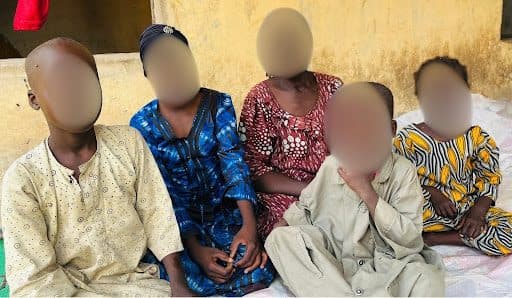 Five children in colorful traditional clothing sit together, against a yellow wall, with faces blurred for privacy.