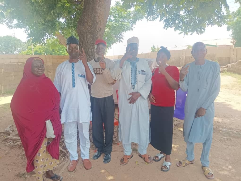 Group of six people standing under a tree, smiling and making hand gestures.