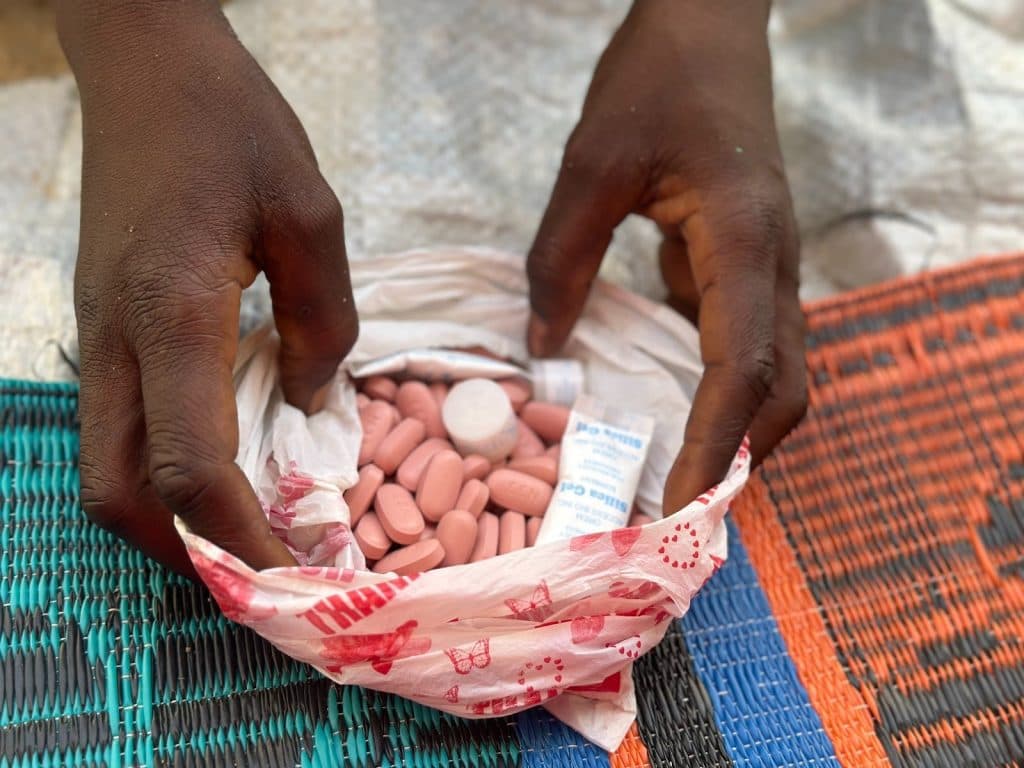 Hands holding a plastic bag filled with pink pills on a woven mat background.