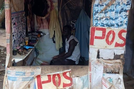 Man sitting in a small makeshift barbershop adorned with haircut posters and "POS" signs.