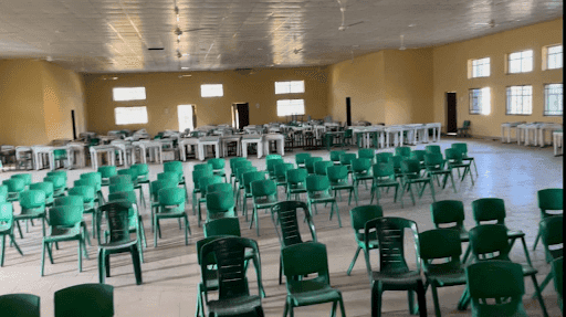 mass abduction in kebbi state, Empty hall with green chairs arranged in rows and tables at the back, featuring beige walls and multiple windows.