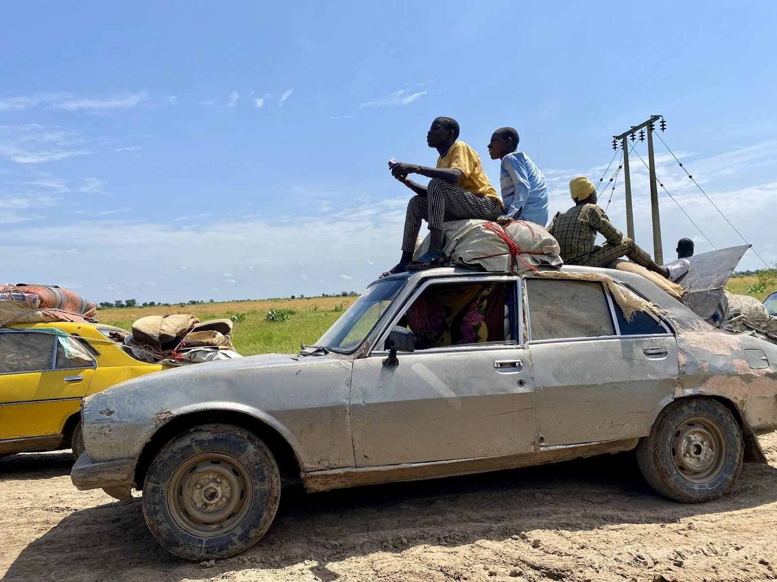 People sit atop a heavily loaded car on a rural road, with fields and power lines in the background under a clear blue sky.