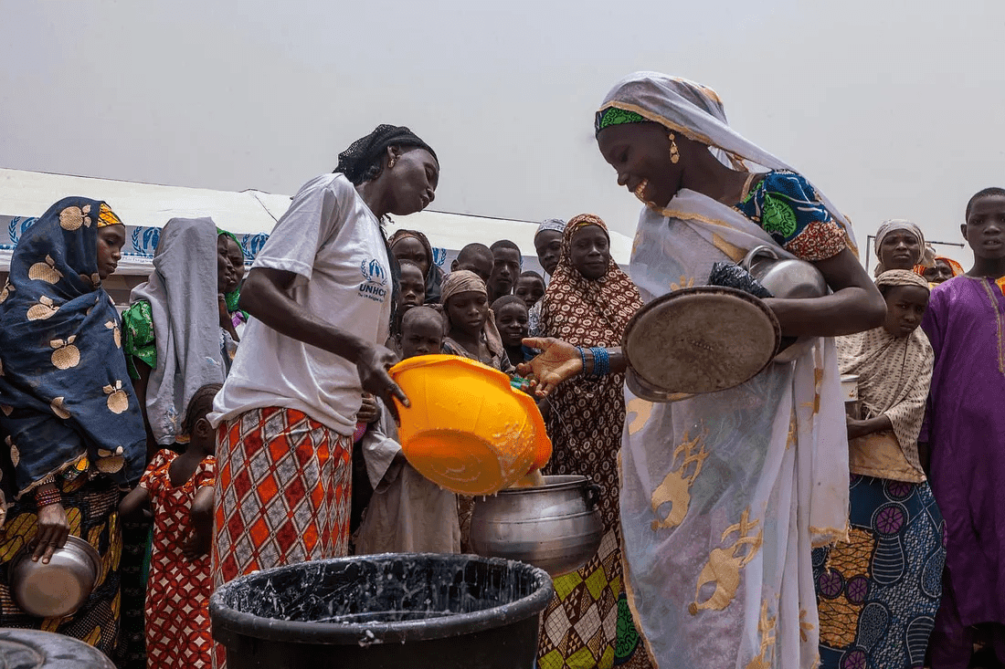 Women and children gather to receive food from a volunteer with a yellow bucket at a distribution event.