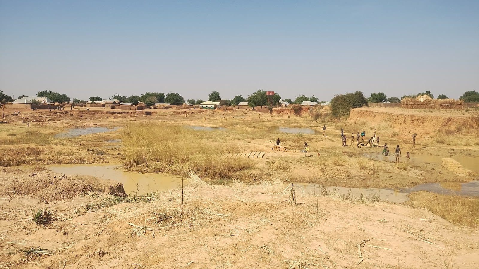 Dry riverbed with children playing, surrounded by mud and sparse vegetation; village huts in the background under a clear blue sky.