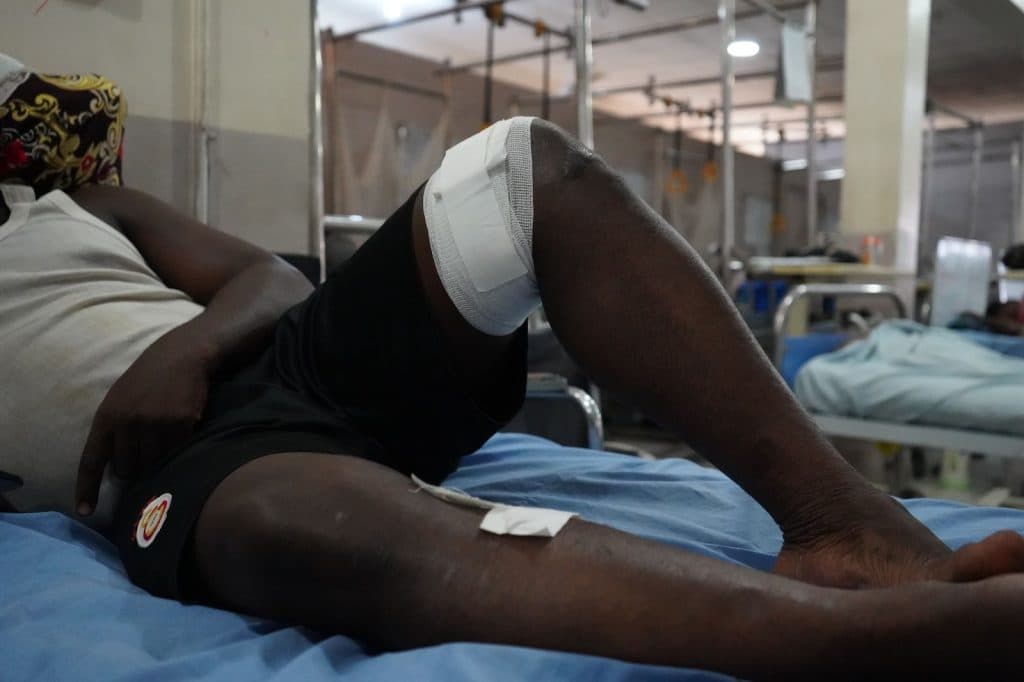Person lying on hospital bed with a bandaged knee in a dimly lit ward.