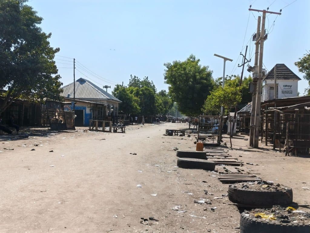 Dusty street with scattered debris, lined with trees and stalls. Sparse activity and bright afternoon sky.